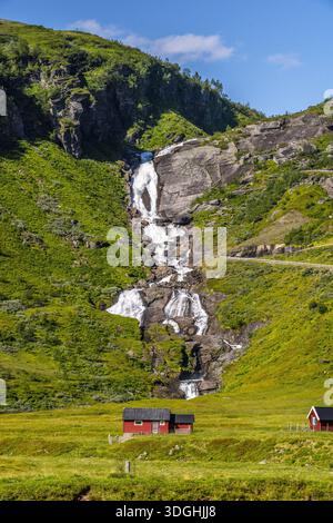 A cascading waterfall flows over green hills next to a red hut in the sunshine Stock Photo