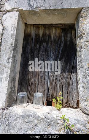 A rustic stone wall frames a weathered wooden window, with ivy and plants growing along the top and sides. Two glass cups rest on a sunlit ledge, evok Stock Photo