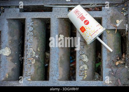 Discarded MacDonalds drinks cup with plastic straw on drain cover at the roadside, MacDonalds drinks carton or container thrown away littering street. Stock Photo