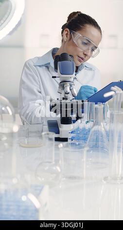 Female scientist working in a laboratory examines bacterial culture samples with a microscope and takes notes on a clipboard. Biotechnology, pharmaceu Stock Photo