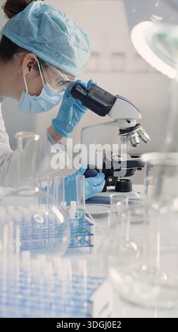 Female scientist wearing mask, safety glasses, gloves and hair cap works in a laboratory using a microscope. Microbiology, biotechnology, medical and Stock Photo