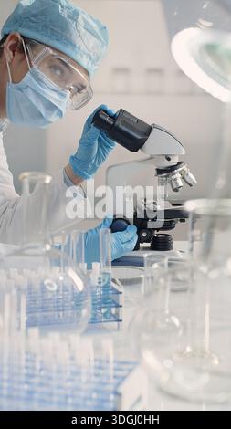 Female scientist wearing mask, safety glasses, gloves and hair cap works in a laboratory using a microscope. Microbiology, biotechnology, medical and Stock Photo