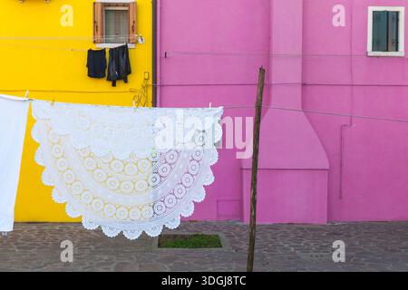 A large, intricately crocheted white tablecloth hangs on a clothesline, drying against vibrant pink and yellow house walls. A concept of traditional c Stock Photo