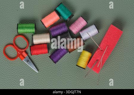 Spools thread in different colors, scissors and sewing needles on green leather background Stock Photo