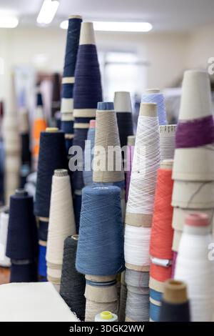 A selective focus view of various colorful yarn cones stacked on a table in a knitwear factory. The bobbins of blue, white, and red thread are ready f Stock Photo