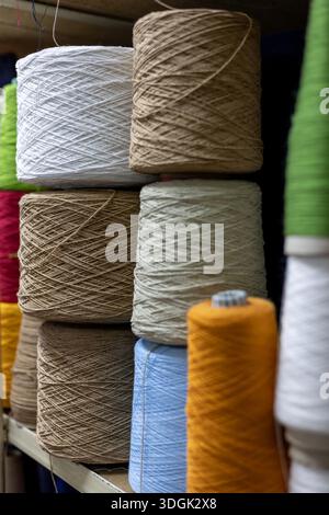 A close-up view of large yarn cones stacked on a shelf in a knitwear factory. The colorful bobbins of white, brown, blue, and orange thread are raw ma Stock Photo