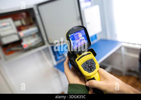 An electrician's hand holds a thermal imaging camera to inspect an open electrical panel, checking for overheating components, hotspots, and potential Stock Photo