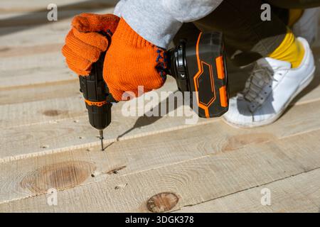 A close-up of a worker's hands in orange gloves using a cordless electric drill to drive a screw into a new wooden deck. This DIY construction project Stock Photo
