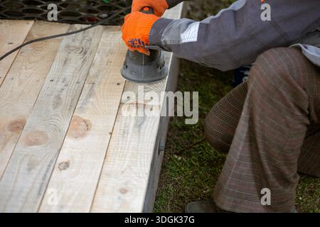 A worker in an orange glove uses an electric orbital sander to smooth new, light-colored wooden planks. This DIY woodworking project involves preparin Stock Photo