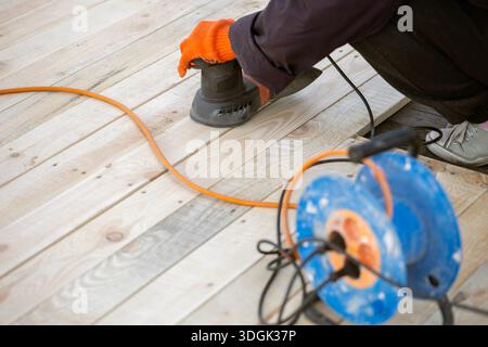 A worker in an orange glove uses an electric orbital sander to smooth a new wooden deck. An orange extension cord and a blue cable reel are on the pla Stock Photo