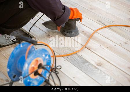 A worker in an orange glove uses an electric orbital sander to smooth a new wooden deck. An orange extension cord and a blue cable reel are on the pla Stock Photo