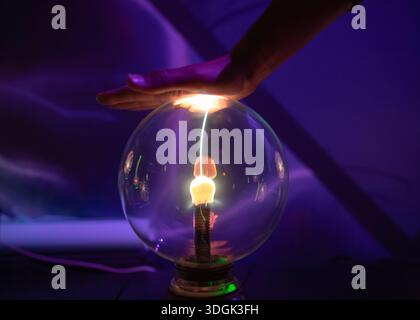 A single, bright filament of electricity jumps from the central electrode of a plasma ball to a hand hovering above, demonstrating static interaction Stock Photo