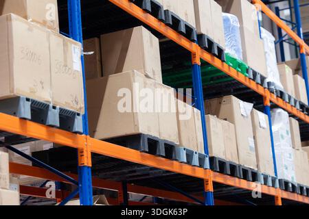 Cardboard boxes are stacked on tall, orange and blue industrial racking in a modern warehouse. The boxes, marked with handwritten numbers, are stored Stock Photo