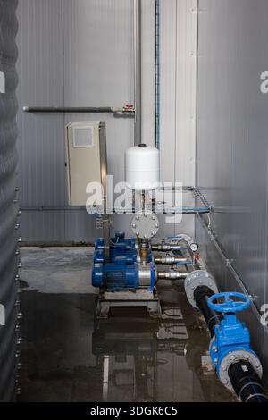 A modern industrial pump station in a factory utility room. A blue electric motor and pump, a white pressure tank, and a control box are connected by Stock Photo