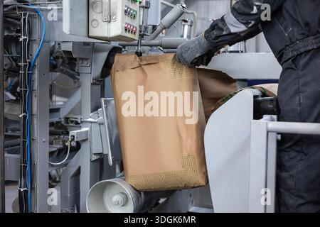 A factory worker in gloves operates an automated bagging machine. The industrial equipment is filling a large brown paper sack with bulk material. The Stock Photo