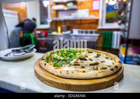 Delicious hot round pizza with two different halves on a wooden board stands on the counter in a busy pizzeria kitchen with chefs working in the blurr Stock Photo