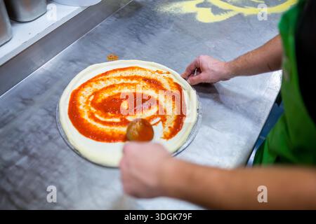 Close up view of a chef hands spreading red tomato sauce on a round raw dough base with a ladle spoon preparing pizza in the professional restaurant k Stock Photo