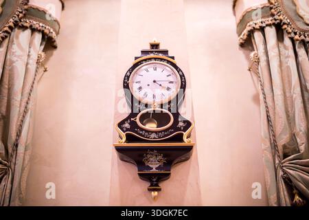 Beautiful antique black wooden wall clock with gold floral decoration and a pendulum hangs on the beige wall between heavy luxurious curtains creating Stock Photo