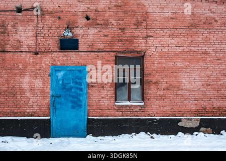 Old weathered red brick wall with a bright blue metal door and a wooden window stands above the white snowy ground in winter creating a colorful vinta Stock Photo
