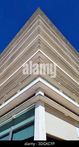 Low-angle architectural photograph capturing the sharp triangular corner of a modern multi-story building against a vivid blue sky, emphasizing geomet Stock Photo