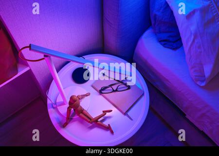 Nightstand featuring a wooden mannequin, notebook, glasses, and a modern lamp illuminating the scene in neon light Stock Photo