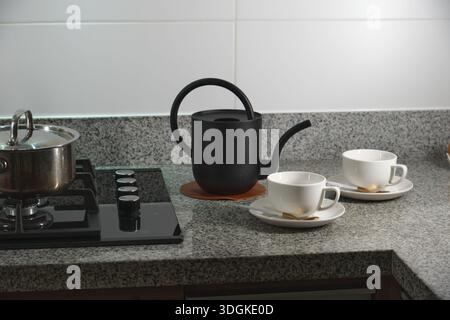 Black teapot and two white ceramic cups resting on granite countertop, ready for serving Stock Photo