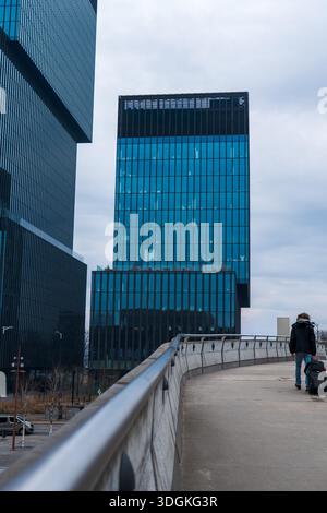 A person walks a curving ramp with a suitcase toward modern glass towers in Katowice, Poland. Stacked offset volumes and vertical window bands stand u Stock Photo
