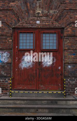 Weathered red double door with barred windows sits in brick arch in Katowice, Poland. White paint patches cover graffiti, hazard stripes mark steps, o Stock Photo