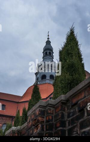 A Baroque dome with clock and cross rises above a red tile roof in Katowice, Silesia, seen from a low angle behind a brick wall and conifers under ove Stock Photo