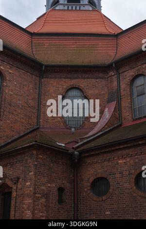 Red brick church facade in Katowice shows an octagonal roof with orange tiles, a quatrefoil window with patterned glass, iron grilles, gutters, and we Stock Photo