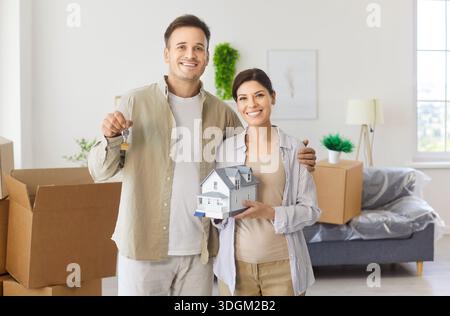 Happy couple holding wooden house model and new keys on moving day at home Stock Photo