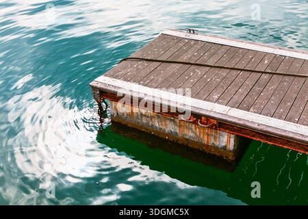 Floating dock on calm green water. Loose rope lies across wooden deck, adding texture to tranquil aquatic scene. Water ripples catching sunlight adjac Stock Photo