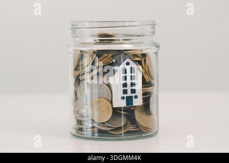 Glass jar filled with coins and a small house model on white background, symbolizing saving for a home, mortgage down payment, real estate investment, Stock Photo