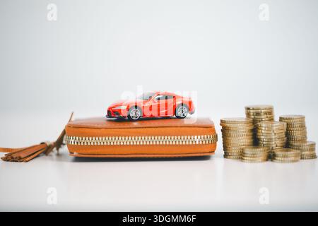 Red toy car on leather wallet beside stacks of coins on white background, symbolizing saving for a vehicle, downpayment, auto loan budgeting, insuranc Stock Photo