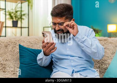 Indian man at home wearing glasses squints reading smartphone text holding screen closer focused. Arabian guy on sofa leans forward eyes narrow tries Stock Photo