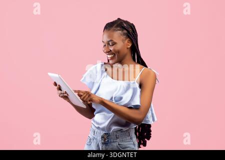 Online life. Pretty young Afro lady using tablet pc for remote work or education on pink studio background Stock Photo