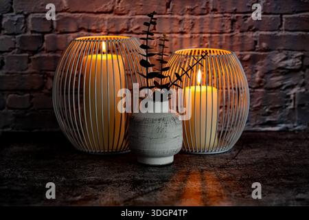 Two candles in two white metal candle holders and artificial leaves in ancient style vase on gray brick wall background. Stock Photo