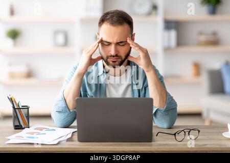Exhausted young man tired after working on laptop for too long, having headache, massaging temples at home Stock Photo