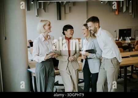Four coworkers are engaged in conversation and sharing laughter in a modern office. They are standing around a table with tablets and laptops present Stock Photo
