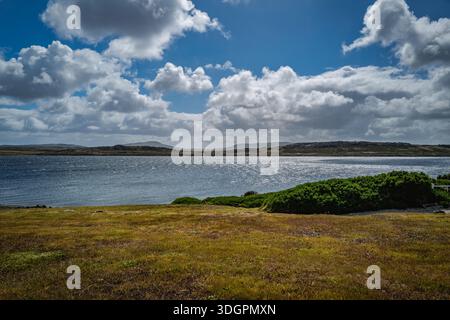 A wide expanse of shimmering water stretches towards distant, rugged hills under a dynamic sky filled with white cumulus clouds Stock Photo