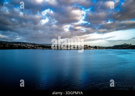 A vast expanse of deep blue water stretches towards a distant city nestled on rolling hills under a dramatic sky filled with dark, textured clouds Stock Photo