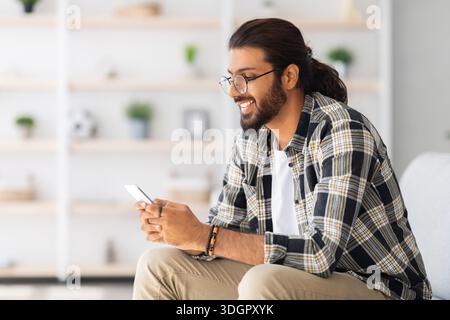 Positive long-haired middle-eastern guy sitting on couch, using cellphone Stock Photo