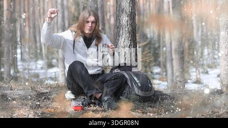 A man in a campaign drinks tea. A young traveler in the forest camped and cooked breakfast in nature. The naturalist drinks hot fresh tea cooked on op Stock Photo