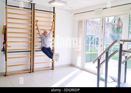 Senior man doing pull ups on wall bars in a rehab center for strength training and physical therapy. This indoor exercise is part of his rehabilitation and recovery process to regain strength and mobility. || Model released Stock Photo