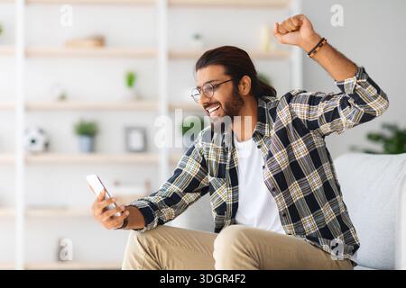 Emotional long-haired indian guy hipster sitting on couch, using smartphone Stock Photo