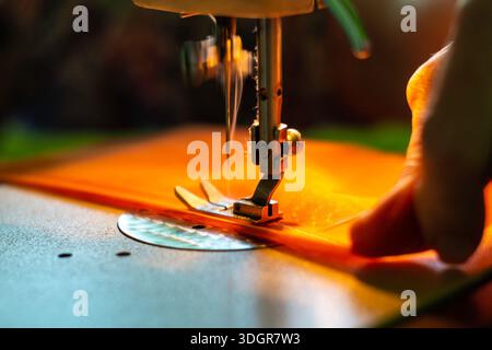 Closeup of Sewing Machine Presser Foot on Orange Fabric with a Right Hand in a Bright Workshop Setting and with Low Depth of Field Stock Photo