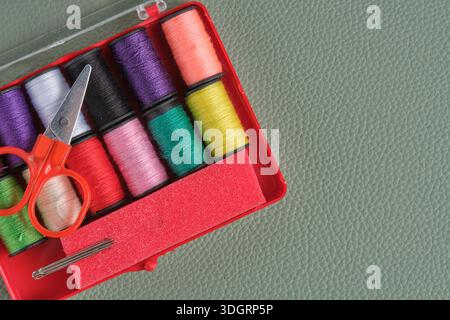 Spools of colorful sewing thread, scissors and needles in red plastic box on green background Stock Photo