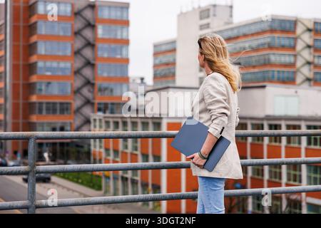 Real estate agent observing city development with laptop. Woman standing in urban area, looking at modern office and industrial buildings Stock Photo