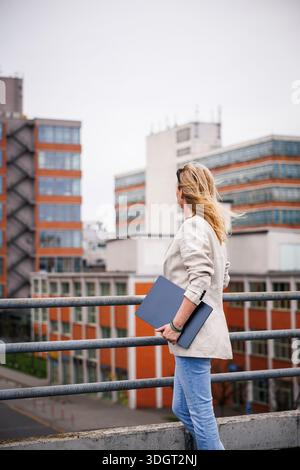 Female real estate developer holding laptop while observing office and industrial buildings in city. Concept of real estate business Stock Photo