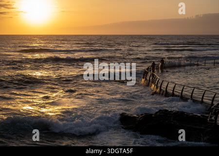 The ocean is calm and the sun is setting. A man is standing on a pier. The water is choppy and the sky is orange Stock Photo